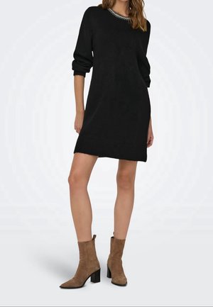 Black knit dress with long sleeves and a round neckline featuring a decorative beaded collar. Model paired with taupe ankle boots.