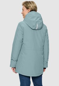 Frau in einer hellblauen Winterjacke mit Kapuze und langen Ärmeln, die seitwärts steht und den Kopf leicht gedreht hat.