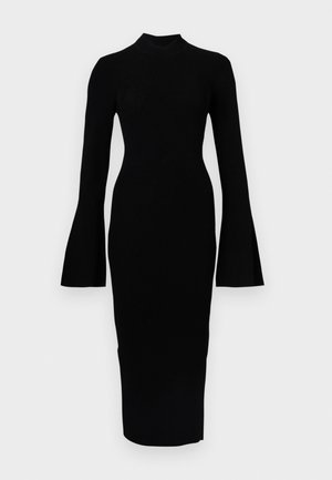 Femme aux cheveux tressés portant une robe midi noire moulante à manches longues et des escarpins slingback noirs à bout pointu, debout devant un fond blanc.