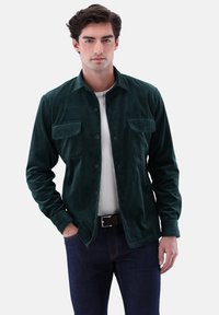 Green corduroy shirt with long sleeves, buttoned front, two chest pockets, and a smooth texture, paired with a white shirt and dark jeans.