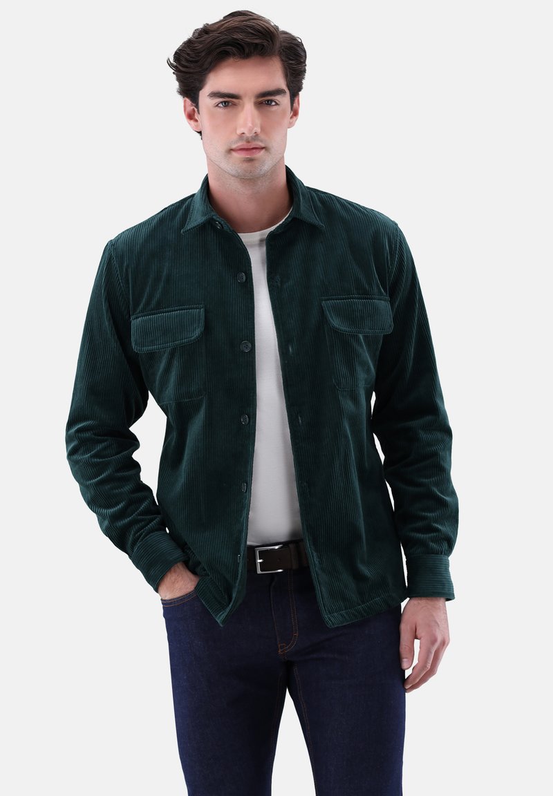 Green corduroy shirt with long sleeves, buttoned front, two chest pockets, and a smooth texture, paired with a white shirt and dark jeans.