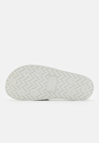 Semelle de basket blanche avec un motif en zigzag texturé, comportant un patch logo proéminent au centre et des grips circulaires pour la traction.