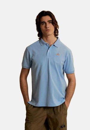 Young man with medium-length dark hair wearing a light blue polo shirt with small logo and brown pants, hands in pockets, neutral background.