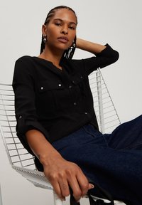 Czarna koszula na guziki z dwiema kieszeniami na piersi, podwiniętymi rękawami i lekką teksturą. Model siedzi na drucianym krześle, ubrany w ciemne dżinsy.