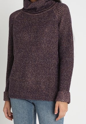 Personne portant un pull à col roulé tricoté violet à manches longues et un jean bleu, debout devant un fond clair uni.