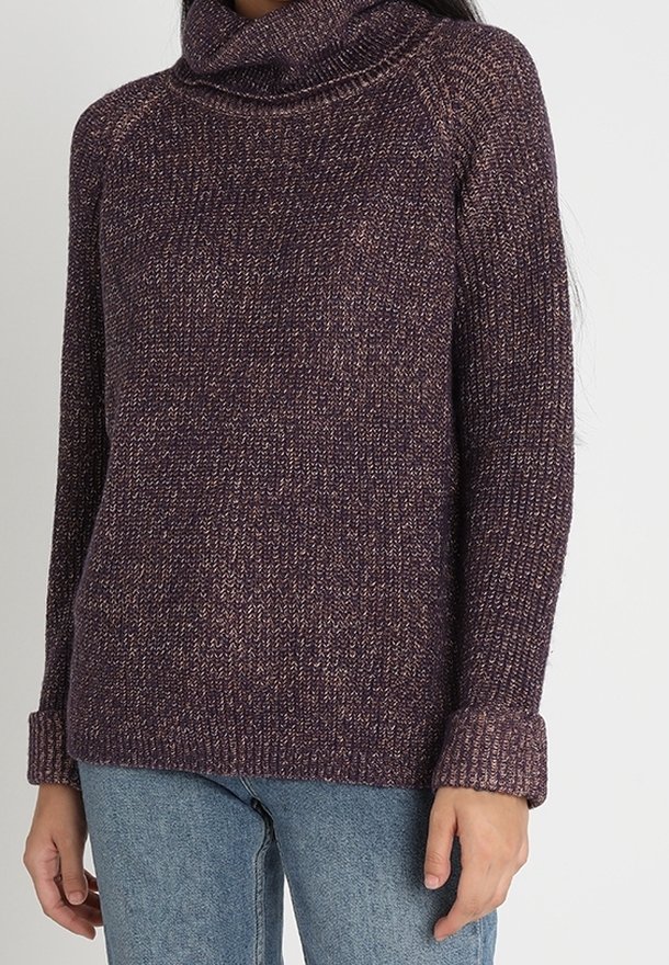 Personne portant un pull à col roulé tricoté violet à manches longues et un jean bleu, debout devant un fond clair uni.