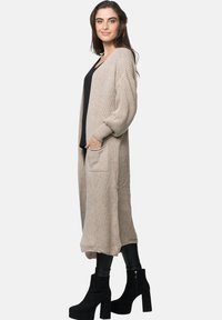 Beige stickad cardigan med långa ärmar, sidfickor och texturmönster, som bärs tillsammans med en svart topp och figursydda byxor, åtföljd av svarta klackar.