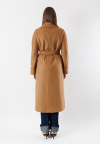 Cappotto lungo in lana color cammello con vita cinturata, colletto a uncino e maniche lunghe, abbinato a jeans scuri con risvolto e tacchi neri.