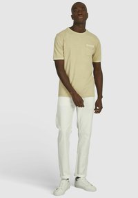 Beige T-shirt met korte mouwen en een borstzak, gecombineerd met witte tapered broeken en witte sneakers, met een neutrale achtergrond.