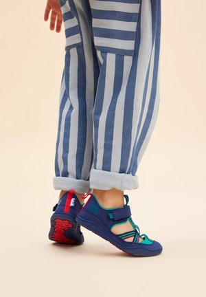 Blue and green children's sandals with adjustable straps, textured soles, and red accents, worn with rolled-up striped pants.