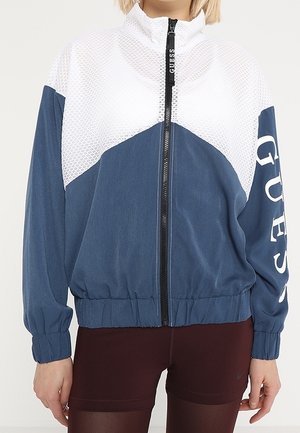 Henkilö, joka käyttää sinivalkoista vetoketjullista takkia verkkokuvioin ja ”GUESS” tekstiä toisessa hihassa, yhdistettynä tummiin leggingsseihin.