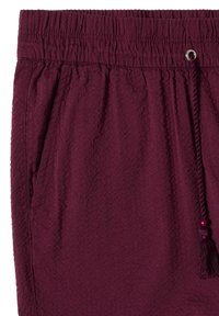 Pantalon texturé bordeaux avec taille élastique et cordon se terminant par un pompon et une perle, montrant le détail de la poche côté droit.