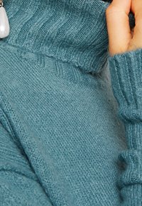 Maglione lavorato a maglia color turchese con colletto e polsini a coste, caratterizzato da una consistenza morbida e una leggera lucentezza, con una mano visibile che tiene il materiale.