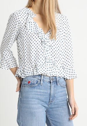 Biała bluzka w granatowe groszki, z falbanką i długością rękawów do łokcia, w połączeniu z jasnoniebieskimi dżinsami z małą czerwoną haftowaną serduszkiem.