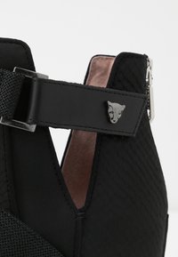 Close-up van een zwarte, gestructureerde schoen met een bandje dat is versierd met een zilveren dierengezicht en een zichtbare rits aan de achterkant.