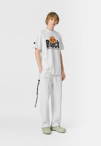 Camiseta blanca de gran tamaño con texto gráfico "Black" y diseño de cara de dibujo animado, combinada con pantalones blancos holgados y calzado texturizado de color verde claro.