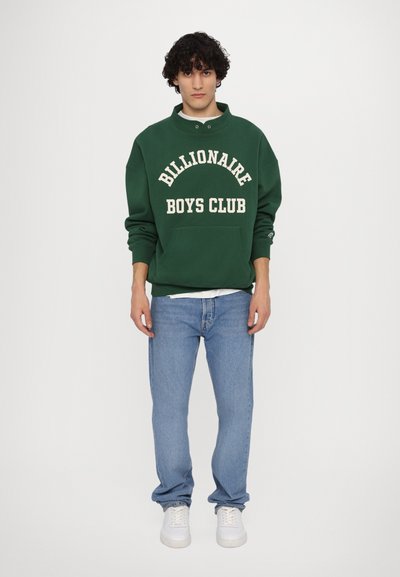 Sweatshirt vert avec "Billionaire Boys Club" en texte blanc, associé à un jean bleu clair et des baskets blanches. Coupe décontractée, design casual.