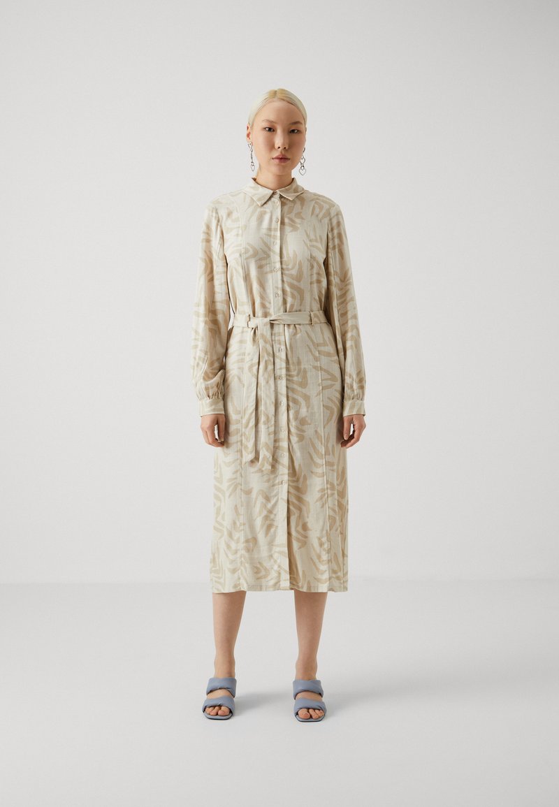 Robe longue beige avec un motif abstrait marron clair, col, fermeture à boutons sur le devant, taille ceinturée et manches longues. Associée à des sandales à glissière bleues.