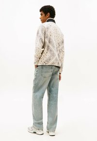 Giacca a fantasia di colore crema con colletto nero, abbinata a jeans blu chiaro a taglio rilassato e sneakers bianche. Presenta design intricati.