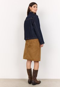 Mörkblå denimjacka med kort design, orange sömmar, bärs över en brun mockakjol. Höga bruna stövlar fulländar outfiten.