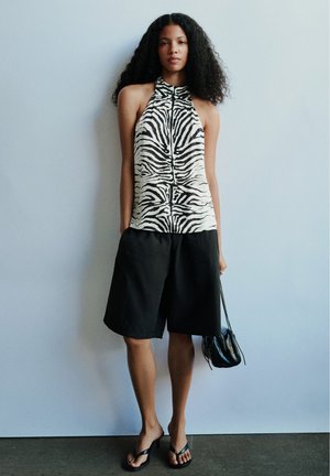 Femme portant un haut sans manches à imprimé zèbre, un short noir longueur genoux, des sandales à talons noires, et tenant un petit sac à main noir.
