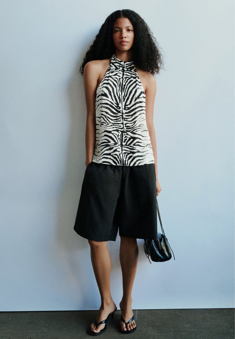 Femme portant un haut sans manches à imprimé zèbre, un short noir longueur genoux, des sandales à talons noires, et tenant un petit sac à main noir.