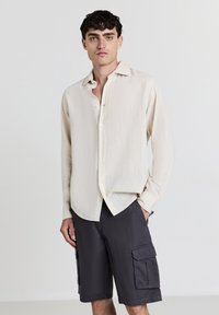 Jeune homme portant une chemise à manches longues beige clair à boutons et un short cargo gris foncé, se tenant debout avec une main dans la poche.