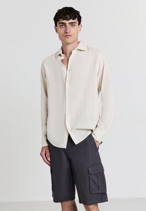 Jeune homme portant une chemise à manches longues beige clair à boutons et un short cargo gris foncé, se tenant debout avec une main dans la poche.