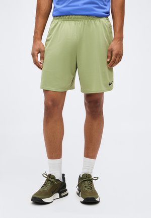 Muž nosi svetlozelené šortky Nike, biele ponožky a olivovo zelené tenisky Nike, stojaci pred jednoliatym svetlým pozadím.