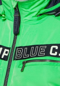 Světlá zelená bunda s černým pruhem a textem "BLUE", se zipovou kapsou a nastavitelnou kapucí se šňůrkami. Hladká, lesklá textura.