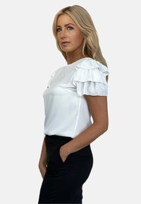 Biała bluzka z krótkim rękawem i falbankami na ramionach, gładka w dotyku, z okrągłym dekoltem, zestawiona z czarnymi spodniami.
