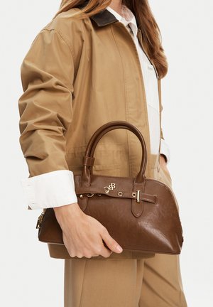 Mujer con chaqueta beige sosteniendo un bolso estructurado de cuero marrón con doble asa y detalles en tono dorado.