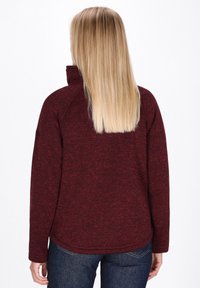 Blondinka obrnjena proč, oblečena v bordo visokokragnast pulover in temno modre kavbojke na enobarvni beli ozadju.