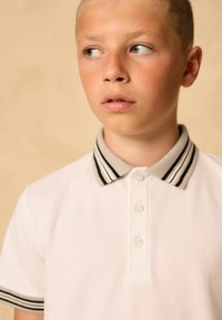 Biała koszulka polo z szaro-czarnym paskiem na kołnierzyku, posiadająca dwa guziki i fakturowany materiał. Design jest klasyczny i prosty.