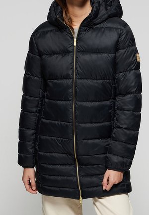 Vrouw draagt een zwarte gewatteerde winterjas met capuchon, gouden voorrits en beige broek, tegen een effen achtergrond.