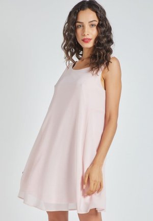 Mujer con cabello rizado vistiendo un vestido de tirantes, suelto y de color rosa pálido, hasta la rodilla, de pie contra un fondo gris claro.
