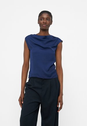 Femme portant un chemisier sans manches bleu marine avec un col drapé et un pantalon noir taille haute à jambes larges, debout devant un fond uni.