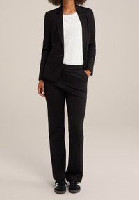 Person klädd i en svart kavaj och byxor, vit T-shirt och svarta sneakers, står med ena handen i fickan mot en beige bakgrund.