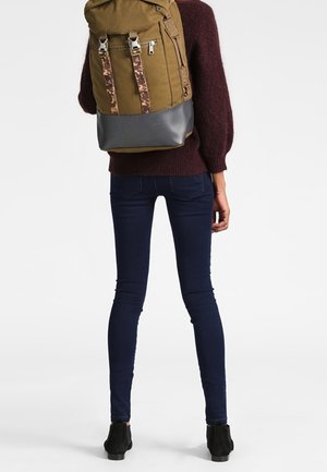 Sac à dos en toile vert olive avec des bretelles à motifs et un fond gris. Porté par une personne en pull bordeaux et jean foncé, de dos.