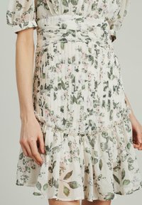 Fließendes, plissiertes Kleid in hellbeige mit floralen Mustern in Grün und Pink. Volantsaum und strukturierten Stoff sorgen für visuelles Interesse.