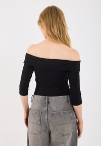 Femeie blondă întoarsă, îmbrăcată cu un top negru off-the-shoulder cu mâneci trei sferturi și blugi gri din denim cu talie înaltă.