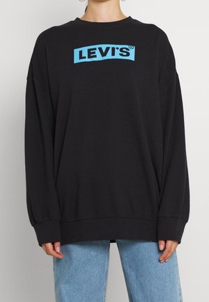 Osoba ubrana w oversize'ową czarną bluzę Levi's z jasnoniebieskim logo i jasnoniebieskie dżinsy na jednolitym tle.