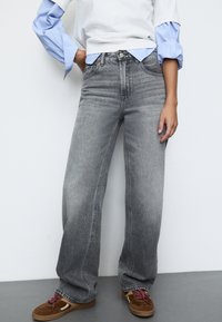 Šedé kalhoty s širokými nohavicemi a vysokým pasem, s pěti kapsami a jemným vyblednutím; doplněné o bílo-modrou pruhovanou košili s dlouhým rukávem.