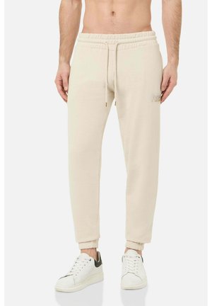 Mann trägt beige Jogginghosen mit elastischem Bund und Bündchen, kombiniert mit weißen Sneakers mit schwarzen Fersenakzenten.