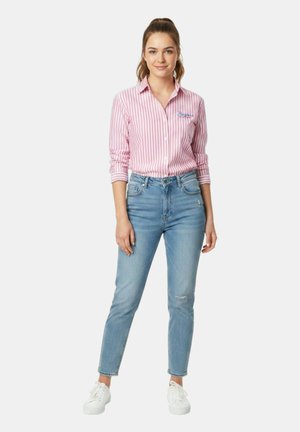 Giovane donna in piedi con una camicia a righe rosa e bianca, jeans blu a vita alta e sneakers bianche, con i capelli legati.