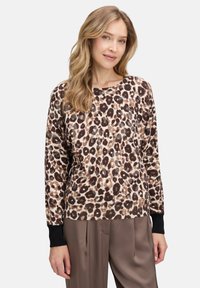 Kobieta z długimi blond włosami, ubrana w sweter w leopardzi wzór z czarnymi mankietami i luźnymi brązowymi spodniami, stojąca na jednolitym tle.