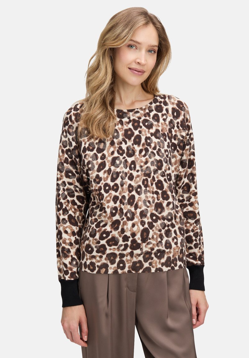 Kobieta z długimi blond włosami, ubrana w sweter w leopardzi wzór z czarnymi mankietami i luźnymi brązowymi spodniami, stojąca na jednolitym tle.