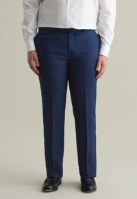 Pantaloni in lino blu navy con taglio su misura, caratterizzati da una vita piatta, passanti per la cintura e pieghe discrete. Abbinati a scarpe eleganti nere.