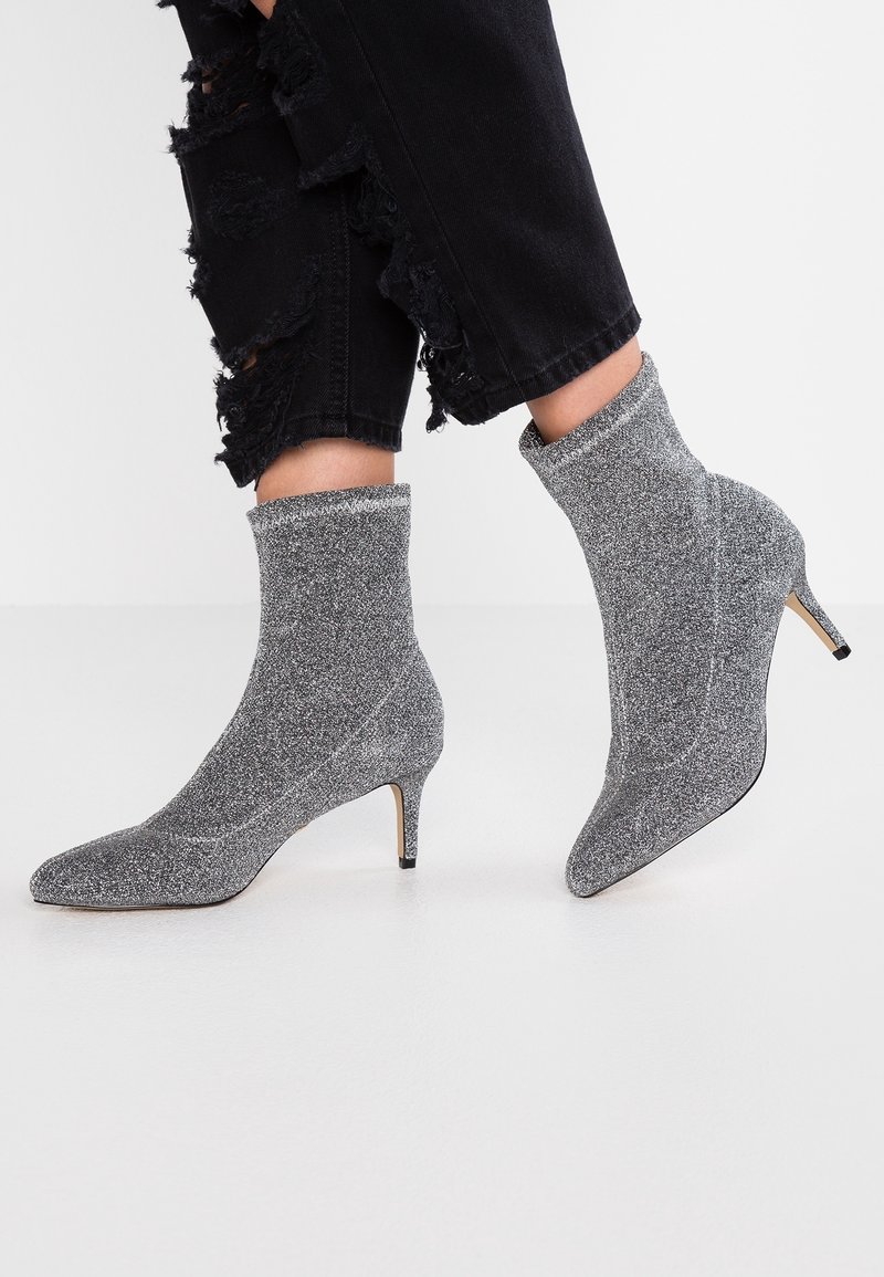Bottines à paillettes argentées avec des bouts pointus et des talons fins. Fabriquées en matériau extensible, elles présentent un col côtelé et une silhouette épurée.