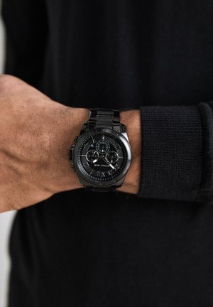 Sort metal håndledsur med kronograf-skiver og romerske tal båret på venstre håndled over sort langærmet.